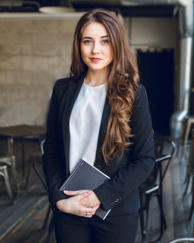 Brunette business woman with wavy long hair and blue eyes stands holding a notebook in hands. She wears a black suit and a white blouse. She is in a cafe with grey walls and dark furniture.